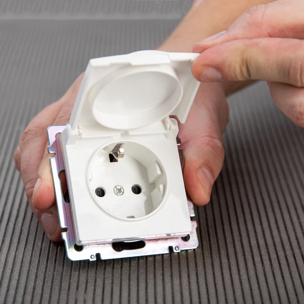 elctrontechnics - Jems Depannage Renovation Une main ouvrant un couvercle de prise électrique étanche de couleur blanche montée sur un cadre métallique, montrant la prise européenne à l'intérieur. La prise est installée sur un fond de tissu rayé gris.