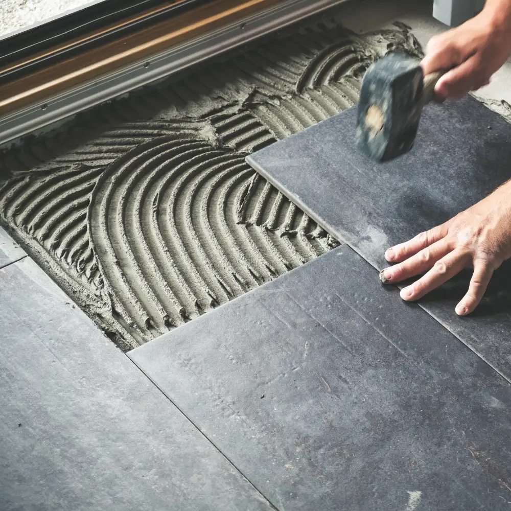 Installation de carreaux de sol gris foncé. Une main place un carreau sur un lit de mortier frais avec un motif de peigne, à côté d'autres carreaux déjà posés et alignés.