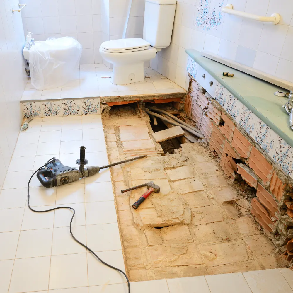 Une salle de bain partiellement rénovée, montrant un sol carrelé intact et un mur partiellement démolis exposant les briques et les tuyaux. Un outil de démolition et un marteau reposent sur le sol à côté d'une perceuse.
