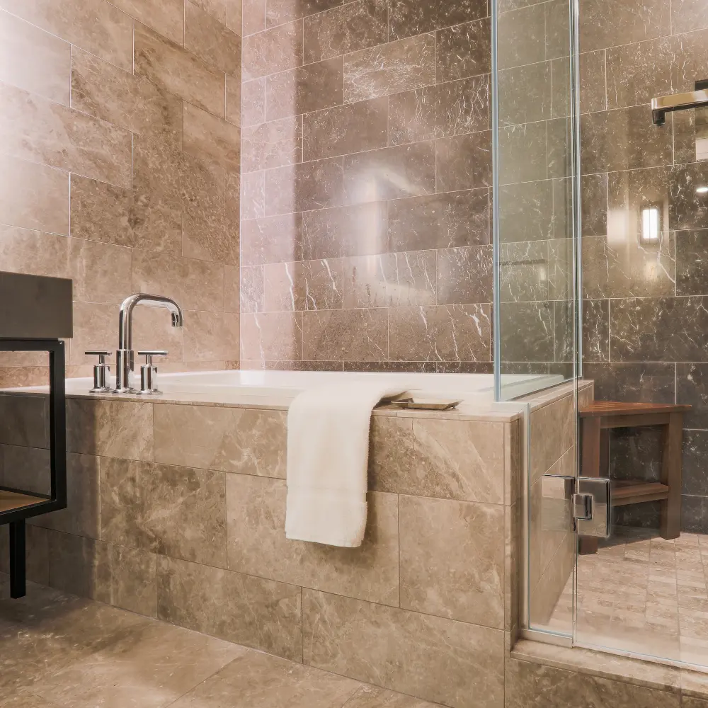 Une salle de bain luxueuse avec des carreaux de marbre sur les murs et un plan de travail, et une douche vitrée. Un banc en bois est placé dans la douche, et une serviette blanche est suspendue au bord de la baignoire.