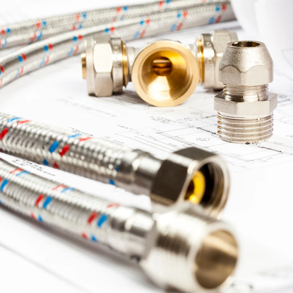 plomberie sanitaire - Jems Depannage Renovation Une sélection de tuyaux flexibles en acier inoxydable et de raccords en laiton sur un fond de plans techniques. Les pièces métalliques brillent et sont disposées en avant-plan, avec le papier en arrière-plan légèrement flou.