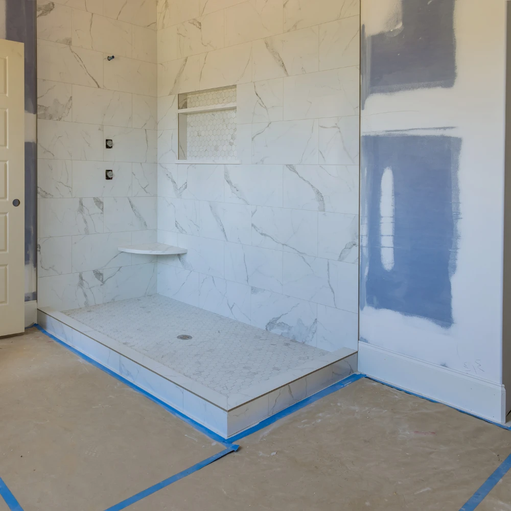 rénovation partiel Une douche à l'italienne en cours de rénovation avec des carreaux de marbre blanc sur les murs et un receveur de douche en mosaïque. Le mur adjacent est à moitié peint et protégé par du ruban de masquage bleu, indiquant les travaux en cours.